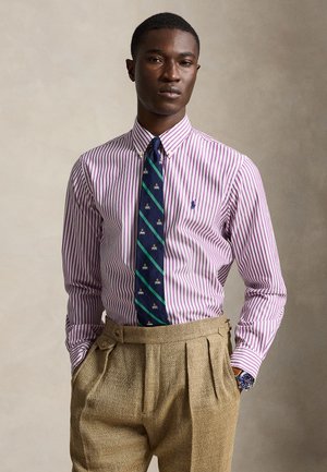Homme portant une chemise de costume violette rayée, une cravate bleu marine à motifs, un pantalon beige plissé et une montre-bracelet, debout les mains dans les poches.