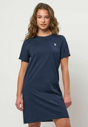 Mladá žena s vlnitými vlasmi, ktorá nosí krátke tmavomodré voľnočasové šaty s malým bielym logom na hrudi, stojaca pred jednoduchým pozadím.