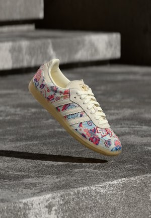 Zapatilla blanca con patrones florales rojos y azules, suela crema y cordones, flotando sobre escalones de cemento gris en un entorno al aire libre.