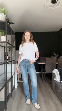 Ανοιχτόχρωμα τζιν παντελόνια με ίσια γραμμή, συνδυασμένα με μια απλή λευκή βαμβακερή μπλούζα και μπεζ σανδάλια. Εσωτερικός χώρος με μινιμαλιστική διακόσμηση.