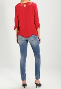 Blusa rossa semi-trasparente con maniche in pizzo a tre quarti, schiena a chiave e orlo ondulato, abbinata a jeans skinny blu chiaro.