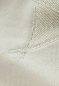 Tela beige claro con una textura suave, que presenta un acento acanalado y costuras visibles. Diseñada con un detalle de costura en forma de V.