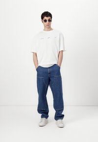 Weißes T-Shirt mit kurzen Ärmeln und blauer Schrift, kombiniert mit lockeren blauen Jeans. Das Modell trägt graue Sonnenbrillen und weiße Sneakers.