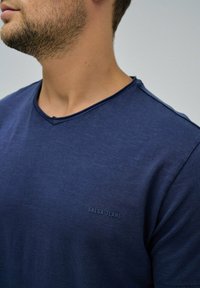Granatowa koszulka typu v-neck wykonana z miękkiej tkaniny, z delikatną fakturą i małym, wytłoczonym logo "Salsa Jeans" na piersi.