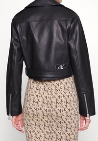 Czarna skórzana krótka kurtka z ząbkowanym kołnierzem, zapinanymi mankietami i małą metką z logo z tyłu. Spódnica ma powtarzający się wzór z logo.