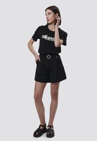 Camiseta negra con el texto plateado "#BOSS", combinada con pantalones cortos negros de tiro alto que cuentan con un cinturón y hebilla redonda. Sandalias negras completan el conjunto.