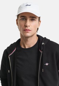 Cappello bianco con logo "Gant", felpa nera con zip e dettagli colorati, e t-shirt nera con un piccolo logo sul petto.