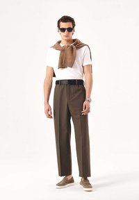T-shirt blanc, pull marron drapé sur les épaules, pantalon marron et chaussures beige sans lacets, complétés par des lunettes de soleil foncées et une montre.