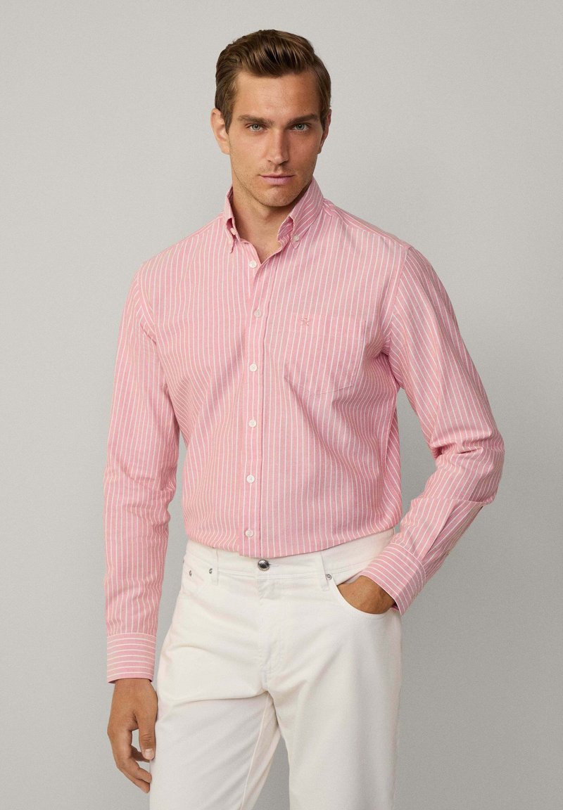 Camicia a maniche lunghe a righe rosa e bianche con colletto, dotata di taschino sul petto e texture di tessuto liscia. Indossata con pantaloni bianchi.
