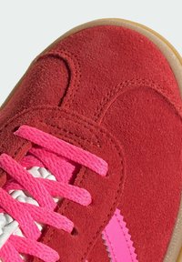 Baskets en daim rouge avec des lacets rose vif et un accent zigzag rose près de la semelle, présentées en gros plan.