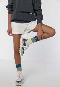 Sneaker sportive con rete grigia, suola bianca e dettagli turchesi; abbinate a calze grigie testurizzate e a righe colorate. Pantaloni corti bianchi e felpa con cappuccio.