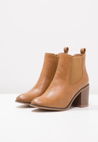 Botas de tobillo de cuero marrón con tacón cuadrado, paneles laterales elásticos y tiradores para ponérselas fácilmente. Textura suave y diseño de punta redonda.