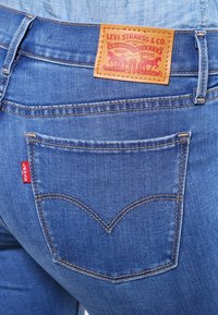 Los vaqueros de mezclilla azul presentan un parche de cuero marrón con la marca en la cintura, y una etiqueta roja en el bolsillo trasero con un diseño de costura distintivo.