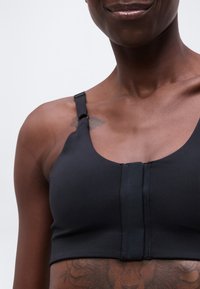 Κοντινό πλάνο ενός ατόμου που φοράει μαύρο αθλητικό σουτιέν με ρυθμιζόμενους ιμάντες και κλείσιμο στο μπροστινό μέρος, δείχνοντας ένα τατουάζ στον ώμο και μέρος ενός τατουάζ στο στήθος.