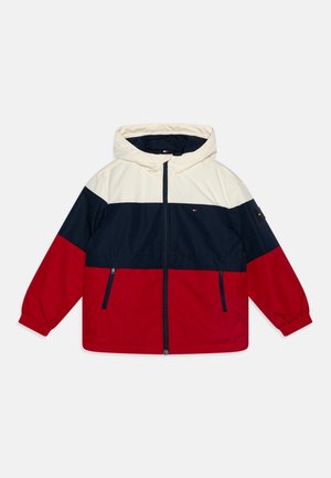 Veste à capuche avec un design en blocs de couleurs crème, marine et rouge. Comprend des poignets élastiques et deux poches latérales zippées.