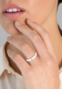 Fehér gyűrű sima felülettel, egy kis arany kiegészítéssel. Középárnyalatú körmökkel viselik, a gyűrű körüli bőr látható.