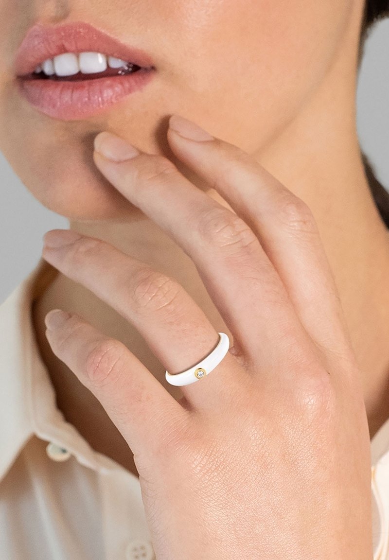 Fehér gyűrű sima felülettel, egy kis arany kiegészítéssel. Középárnyalatú körmökkel viselik, a gyűrű körüli bőr látható.