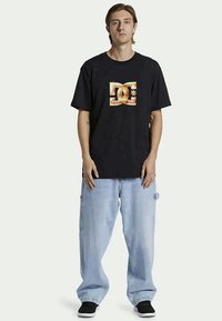 En ung man står, iklädd en svart T-shirt med en gyllene DC-logotyp, ljusblå baggyjeans och svarta sneakers framför en enfärgad bakgrund.