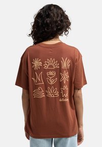 Braunes Baumwoll-T-Shirt mit botanischen und Wellenlinienzeichnungen in cremefarbe auf dem Rücken. Kurze Ärmel, lockere Passform, Rundhalsausschnitt.