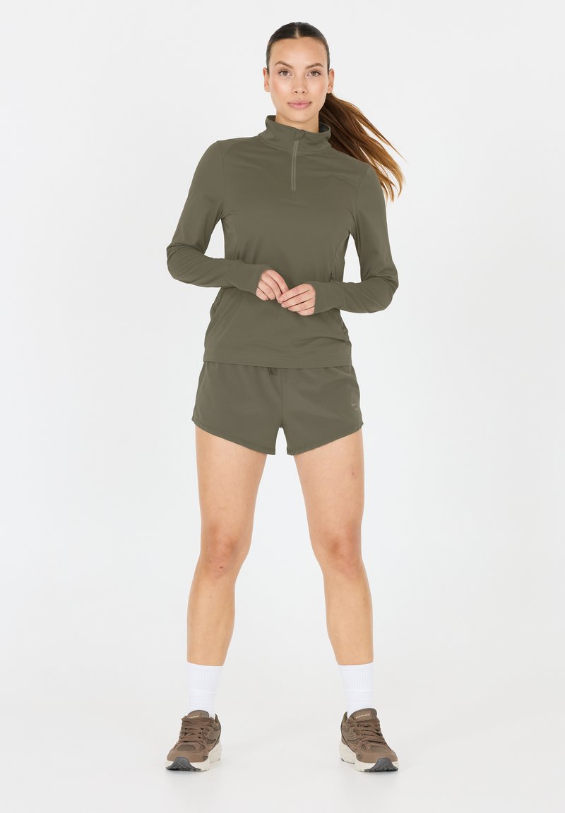 Olivgrüner Langarm-Pullover mit halben Reißverschluss und passender Shorts. Weicher Stoff, sportliche Passform, kombiniert mit hellbraunen Laufschuhen und weißen Socken.