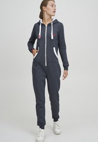 Navy hette-jumpsuit med hvite snorer og lommer. Glatt tekstur, tettsittende passform, og ribbede mansjetter. Rød aksentlogo nær brystet.