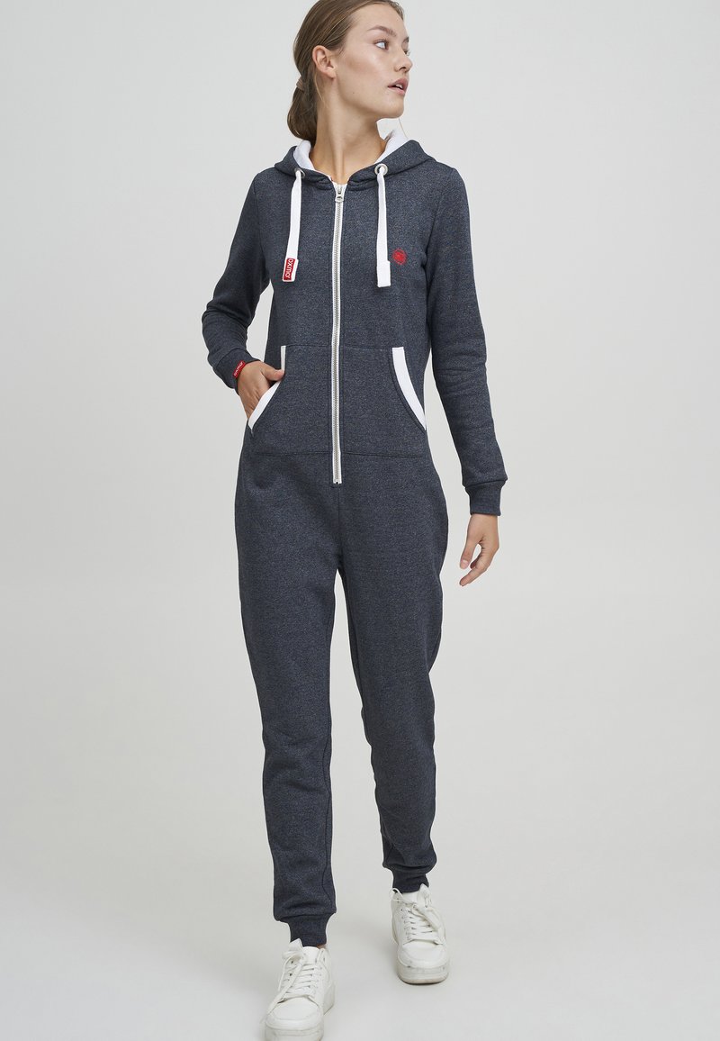 Navy hette-jumpsuit med hvite snorer og lommer. Glatt tekstur, tettsittende passform, og ribbede mansjetter. Rød aksentlogo nær brystet.