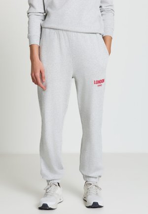 Personne portant un pantalon de survêtement gris clair avec l'inscription "LONDON 1990" en rouge sur la cuisse, des baskets blanches, et un sweat-shirt gris clair, debout les mains dans les poches.