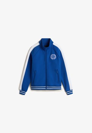 Blue athletic jacket with white stripes, zip closure, two zippered pockets, and a round logo detail on the chest. Soft fabric texture.