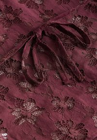 Burgunderfarvet blonderstof med blomsterprint og en tekstureret baggrund. Inkluderer bindebånd til fastgørelse, som fremhæver dets designdetalje.