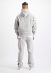 Man met zijn rug naar de camera, gekleed in een lichtgrijze hoodie, bijpassende sweatpants, witte sneakers en een wit shirt onder de hoodie.