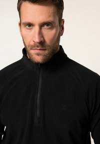 Schwarze Fleecejacke mit halb durchgehendem Reißverschluss, hoher Kragen und dezentes Logo-Detail auf der Brust. Glatte Textur, sportliche Passform.