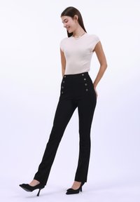 Pantaloni neri a vita alta con dettagli in bottone dorato, abbinati a un top aderente ribbed beige chiaro. La modella indossa tacchi neri.