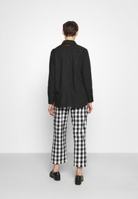 Camicia nera a maniche lunghe con taglio dritto, abbinata a pantaloni a gamba larga a scacchi bianchi e neri. Sfondo semplice e scarpe nere.
