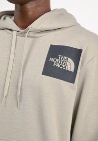 Gros plan d'une personne portant un sweat à capuche beige avec un logo carré noir et le texte « THE NORTH FACE » sur la poitrine.