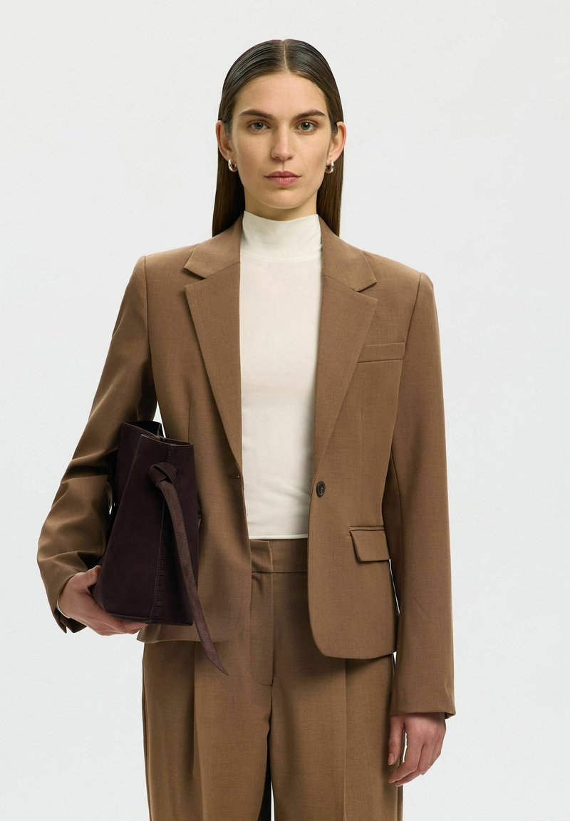 Femme aux cheveux bruns raides porte un costume marron et un haut blanc, tenant un sac à main marron foncé dans sa main gauche sur un fond uni.