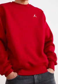 Person wearing a red sweatshirt with a small white Jumpman logo on the chest, hands in pockets, paired with dark gray pants.