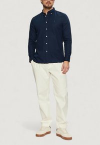 Camicia blu con bottoni, maniche lunghe, pantaloni bianchi e sneakers chiare. La camicia presenta un colletto e un piccolo logo. Tessuto di consistenza liscia.