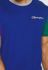 Tričko s krátkým rukávem v modré barvě s červenými a zelenými detaily. Má šedý žebrovaný límeček a malé logo na hrudi. Měkký bavlněný materiál.