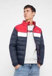 Polstret jakke med røde, hvide og marine farveblokke, høj krave, lynlås foran og elastiske manchetter. Båret over en grå sweatshirt og blå jeans.