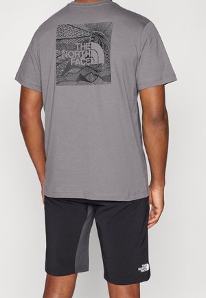 Homme portant un t-shirt gris The North Face avec un motif noir et un short noir avec un logo blanc, debout tourné dos à la caméra.