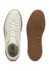 Crèmekleurige leren sneakers met een geperforeerd zijpaneel, witte veters en een bruine rubberen zool met een golvend loopvlakpatroon. Geaccentueerd met een QR-code.