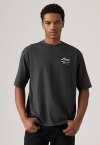 Camiseta de algodón gris oscuro con mangas cortas, cuello redondo y un logo blanco de Levi's en el lado izquierdo del pecho. El modelo posa con las manos en los bolsillos.