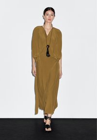 Robe vert olive à manches longues avec un design croisé, présentant un décolleté en V profond et une coupe ample. Un collier à pompons noir et des sandales plates complètent le look.