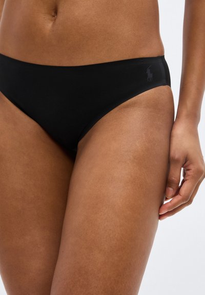 Κοντινό πλάνο ενός ατόμου που φοράει μαύρο κάτω μέρος μαγιό μπικίνι με ένα μικρό κεντημένο λογότυπο στο αριστερό ισχίο, πάνω σε απλό φόντο.