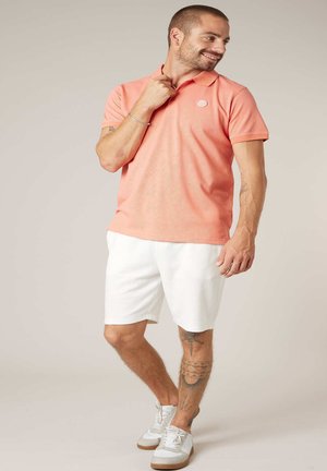 Homme souriant portant un polo corail, un short blanc et des baskets blanches, debout avec une main près du col et des tatouages visibles sur la jambe.