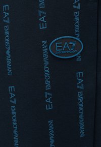 Ciemna tkanina z powtarzającymi się pionowymi logo "EA7 Emporio Armani" w kolorze niebieskim oraz niebieską owalną naszywką EA7 Emporio Armani.