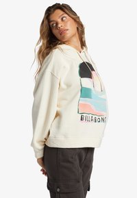 Felpa color crema con un design grafico colorato sul davanti, dotata di cappuccio con cordini e vestibilità rilassata. Abbinata a pantaloni cargo scuri.