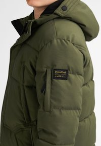 Olivgrüne Pufferjacke mit Kapuze, strukturiertem Obermaterial, zwei seitlichen Taschen und einem Logo-Patch am Oberarm.