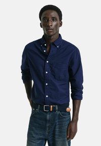 Camicia button-up blu navy con maniche lunghe e tasca sul petto, abbinata a jeans di denim e una cintura intrecciata con un accento in pelle marrone.
