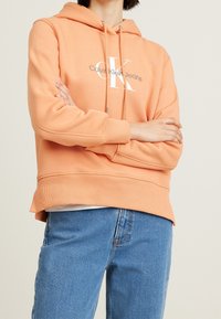 Peach-colored hoodie with a large white "CK" logo and drawstrings. Ribbed cuffs and hem, with a relaxed fit and soft texture. Worn with blue jeans.